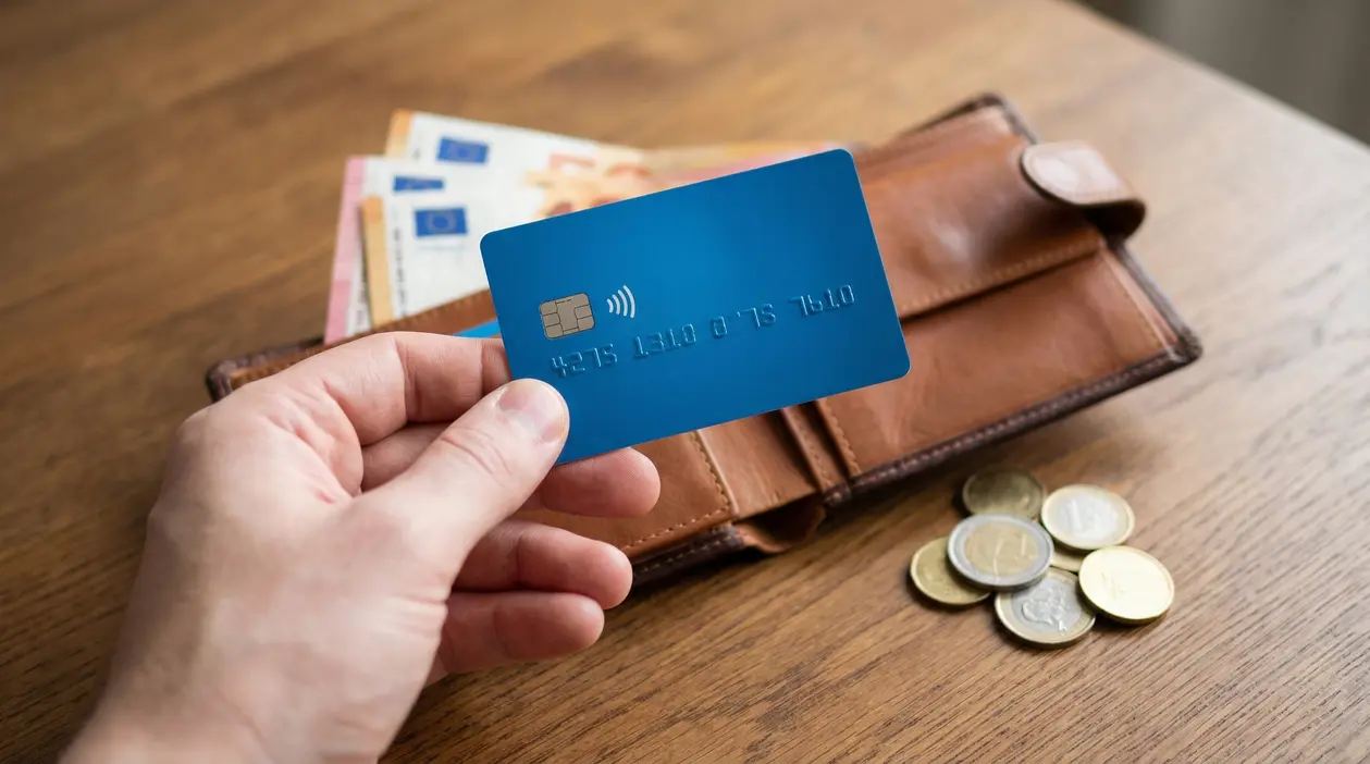 Una mano tiene una carta prepagata blu davanti a un portafoglio con contanti e monete su un tavolo