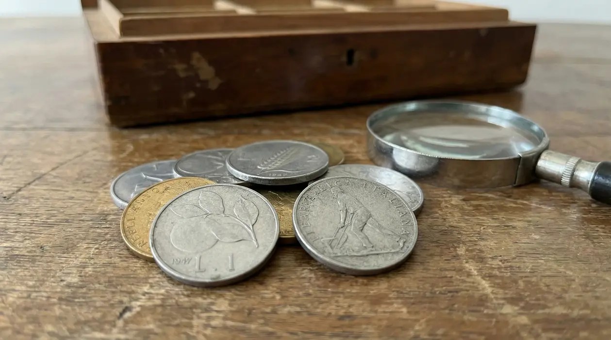Vecchie monete italiane, tra cui una lira del 1947, su un tavolo con lente d'ingrandimento
