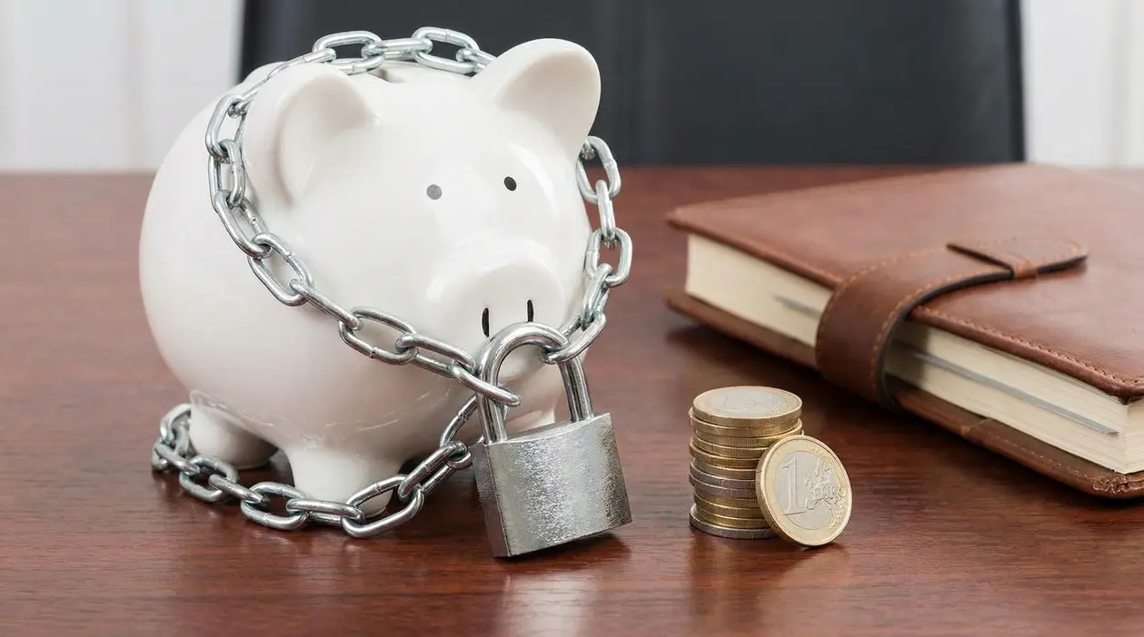 Salvadanaio bianco con catena e lucchetto accanto a monete e taccuino su scrivania