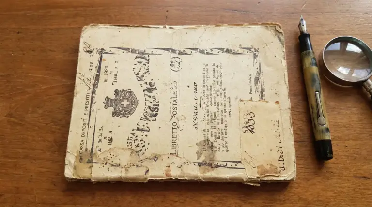 Vecchio libretto postale consunto accanto a una penna stilografica e a una lente d’ingrandimento su un tavolo in legno