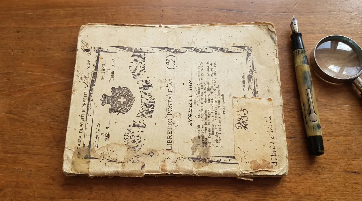 Vecchio libretto postale consunto accanto a una penna stilografica e a una lente d’ingrandimento su un tavolo in legno